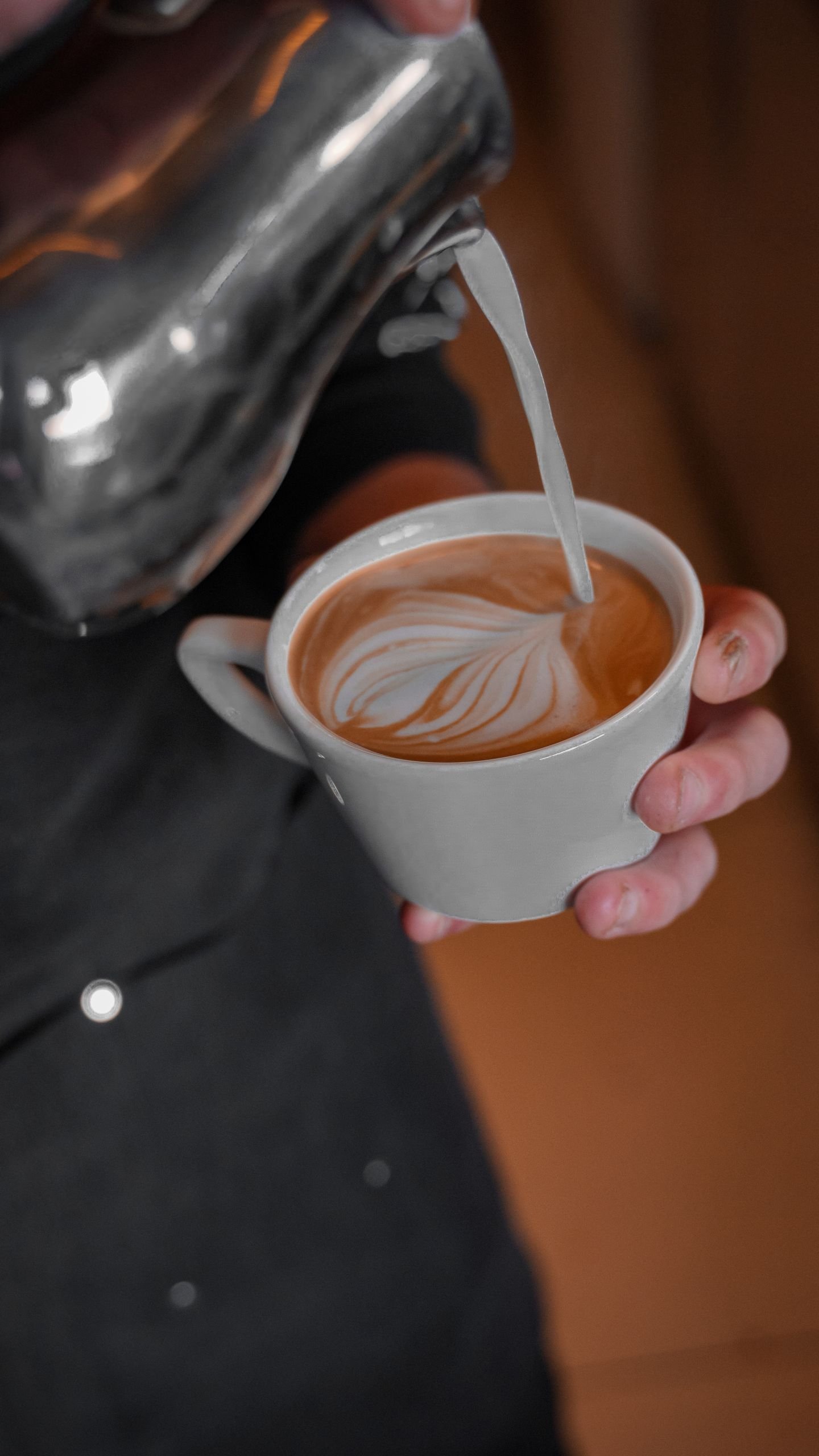 Close-up of hands pouring frothy milk into coffee cup creating intricate latte art.